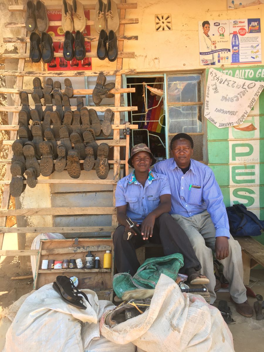 22 year old cobbler Boars (left) sits with his community health worker Stanislaus outside his shop. 