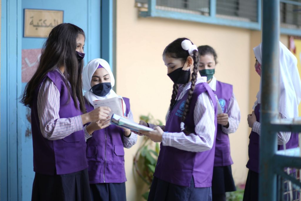Five school children wearing purple uniforms and face masks stand together outside a classroom, reading and talking. They are wearing face masks
