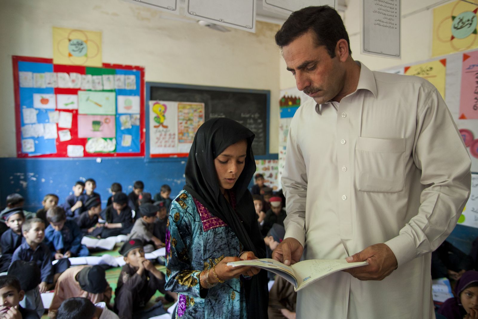 A child and teacher stand at the front of a classroom, the child is reading aloud