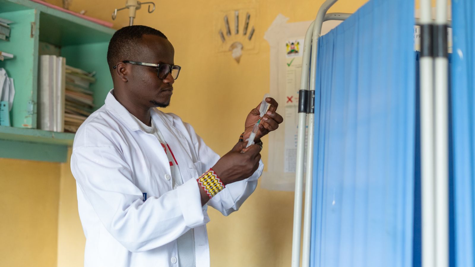 A black man in a lab coat wearing glasses, holds a syringe 