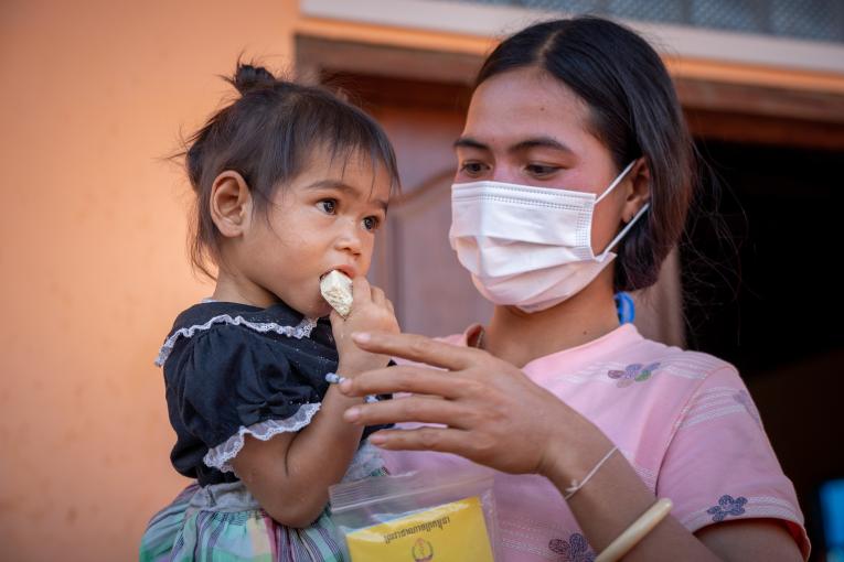 A masked healthcare worker in Cambodia holds a young child eating an emergency ration. 